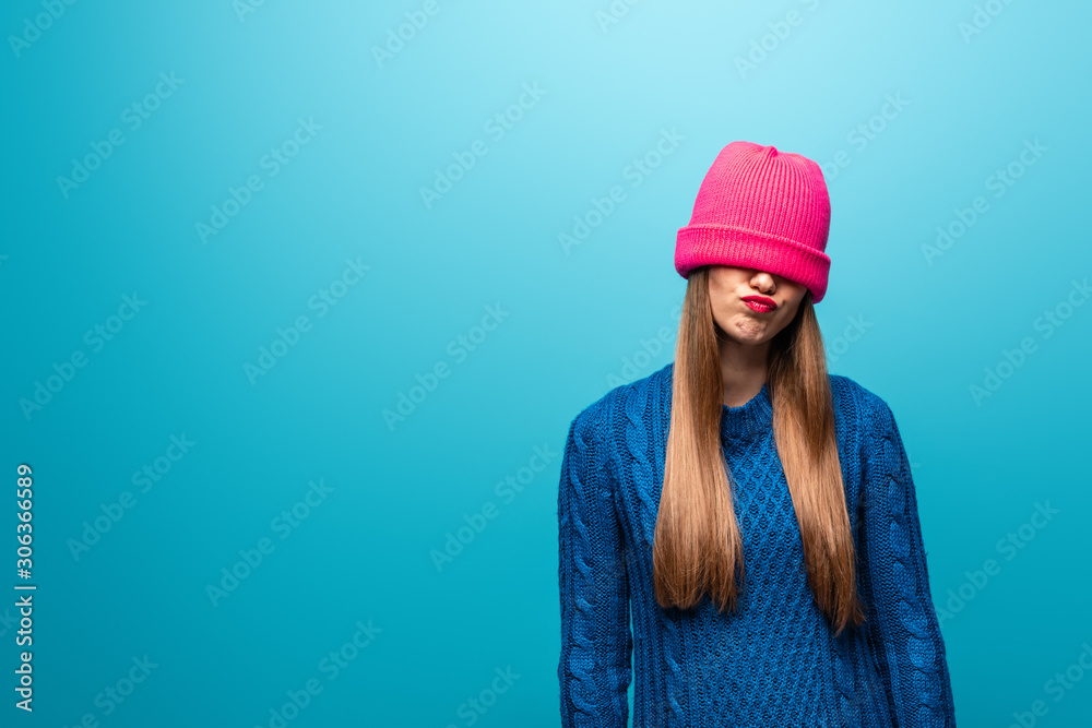attractive offended woman in knitted sweater with pink hat on eyes, isolated on blue
