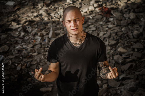 Young brutal bald tattooed guy with both hands shows his middle fingers. A handsome guy shows rude gestures with his fingers against the backdrop of the ruins of an abandoned building.