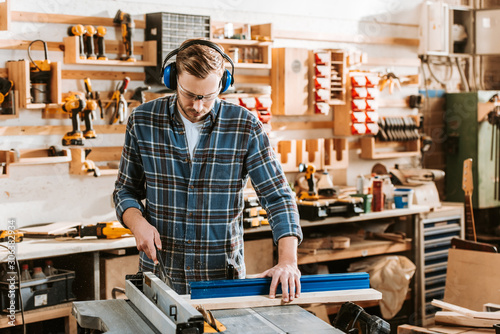 handsome woodworker in protective headphones holding plank near circular saw photo