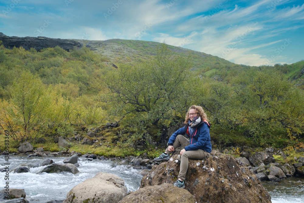 young woman sits on a rock in a fast flowing river, with a blue sky in autumn colours