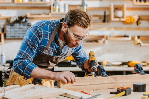selective focus of carpenter in safety glasses and apron holding hammer drill and blowing on sawdust near wooden plank photo