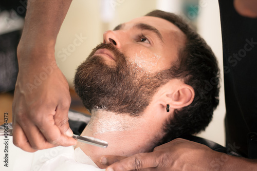 Close-up view of the face of a young man shaving his beard with a blade in the barbershop