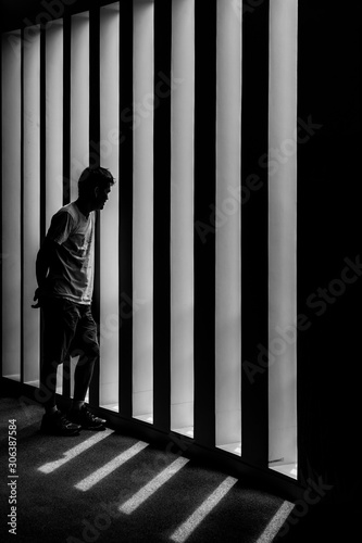 The asian man standing in the dark room with stress moment and he looking outside in the leading line of black and white pattern of light and shadow.