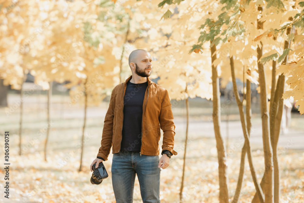 Bald brutal photographer with a beard in a suede leather jacket, blue shirt and jeans poses with the camera in the park in the afternoon