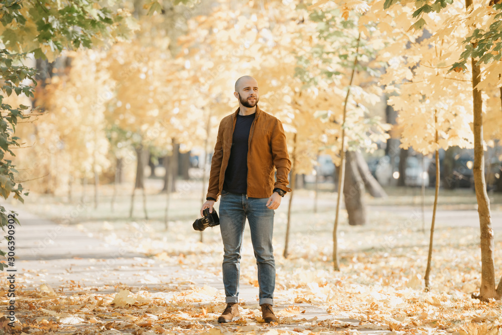 Bald brutal photographer with a beard in a suede leather jacket, blue shirt, jeans, and Chelsea boots looks for somebody in the park in the afternoon Bald brutal photographer with a beard in a suede leather jacket, blue shirt, jeans, and Chelsea boots looks for somebody in the park in the afternoon