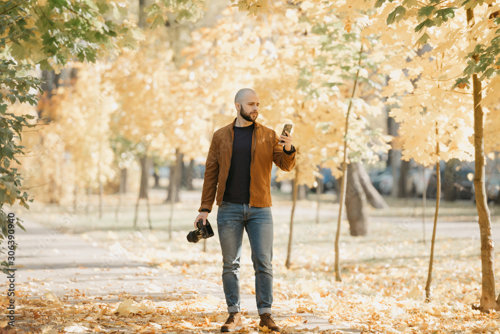 Bald brutal photographer with a beard in a suede leather jacket, blue shirt, jeans, and Chelsea boots holds a camera and a smartphone in the park in the afternoon Bald brutal photographer with a beard in a suede leather jacket, blue shirt, jeans, and Chelsea boots holds a camera and a smartphone in the park in the afternoon
