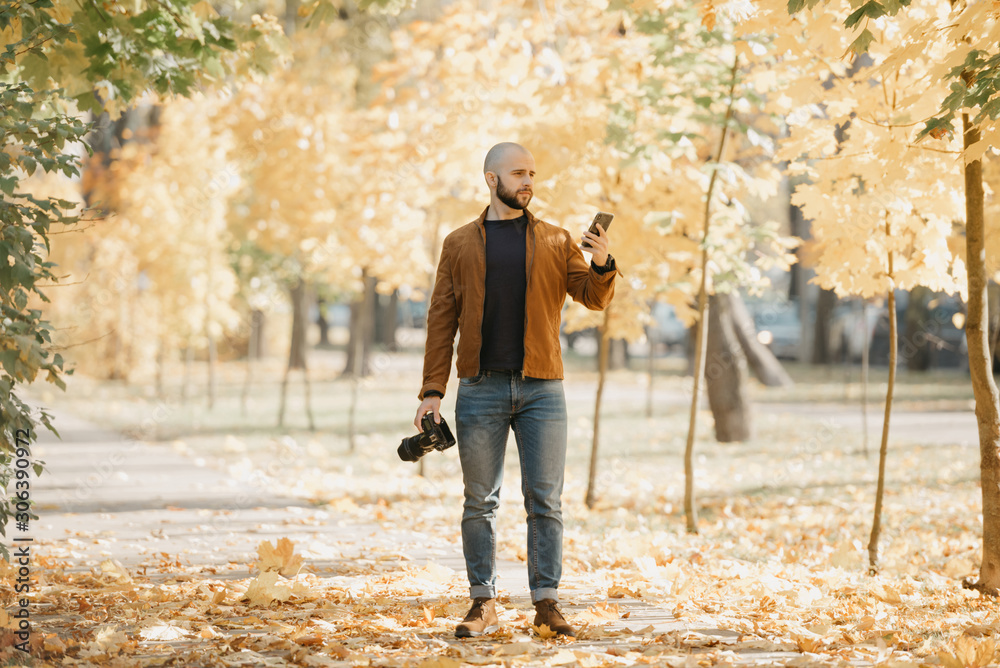 Bald brutal photographer with a beard in a suede brown leather jacket, blue shirt, jeans, and Chelsea boots holds a camera and a smartphone in the park in the afternoon Bald brutal photographer with a beard in a suede brown leather jacket, blue shirt, jeans, and Chelsea boots holds a camera and a smartphone in the park in the afternoon