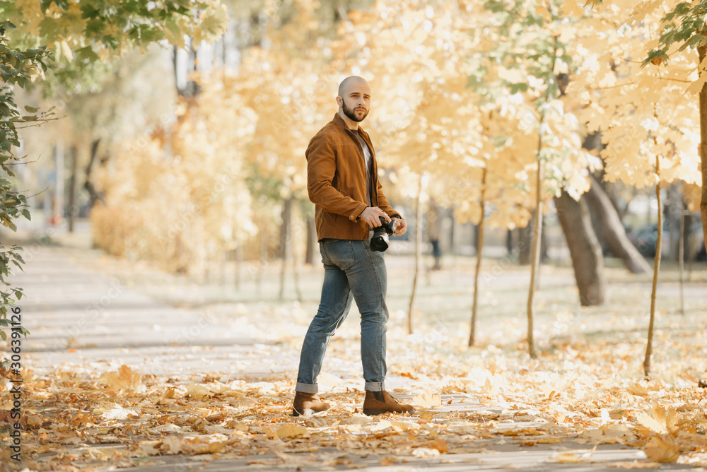 Bald brutal half-turn photographer with a beard in a suede leather jacket, blue shirt, jeans, and Chelsea boots holds the camera and waits for a model in the park in the afternoon