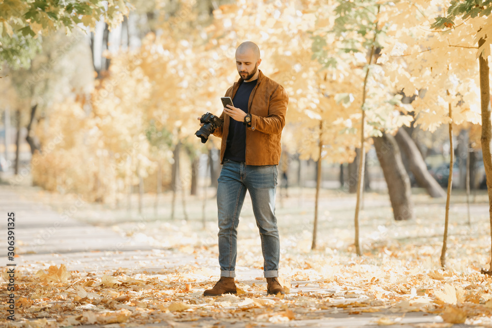 Bald stylish photographer with a beard in a suede leather jacket, blue shirt, jeans, and Chelsea boots holds the camera and the smartphone looks for someone in the park in the afternoon Bald stylish photographer with a beard in a suede leather jacket, blue shirt, jeans, and Chelsea boots holds the camera and the smartphone looks for someone in the park in the afternoon