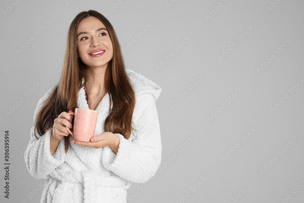 Beautiful young woman in bathrobe with cup of morning drink on light grey background, space for text Beautiful young woman in bathrobe with cup of morning drink on light grey background, space for text