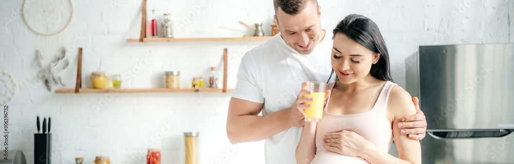 panoramic shot of happy pregnant couple hugging and holding orange juice in kitchen panoramic shot of happy pregnant couple hugging and holding orange juice in kitchen