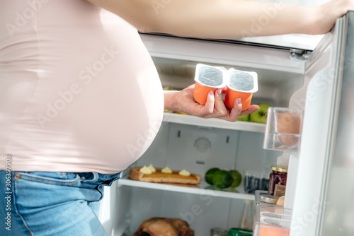 cropped view of pregnant woman holding yogurt near opened fridge in kitchen