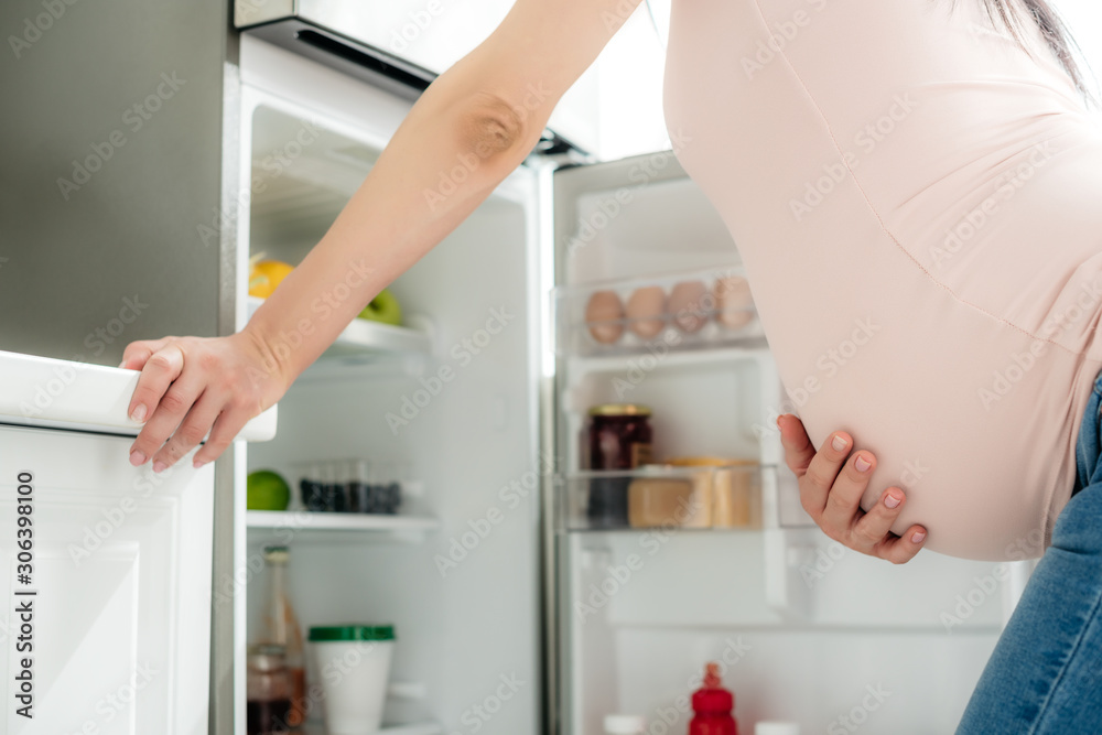 cropped view of pregnant woman holding belly and looking into opened fridge in kitchen cropped view of pregnant woman holding belly and looking into opened fridge in kitchen