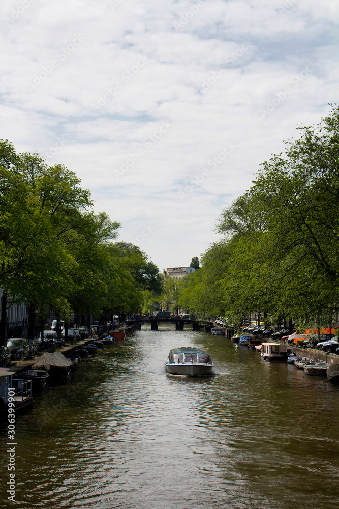 View of a cruise canal tour boat, trees, parked cars and boats in Amsterdam. It is a sunny summer day.