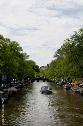 View of a cruise canal tour boat, trees, parked cars and boats in Amsterdam. It is a sunny summer day.