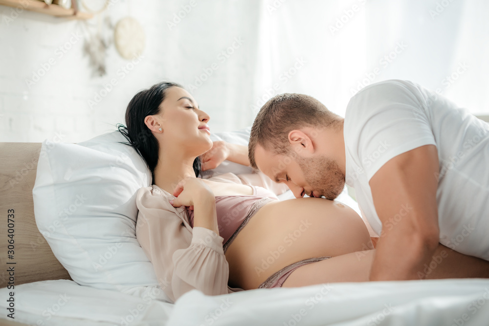 happy husband hugging and kissing tummy of his smiling pregnant wife in bed happy husband hugging and kissing tummy of his smiling pregnant wife in bed