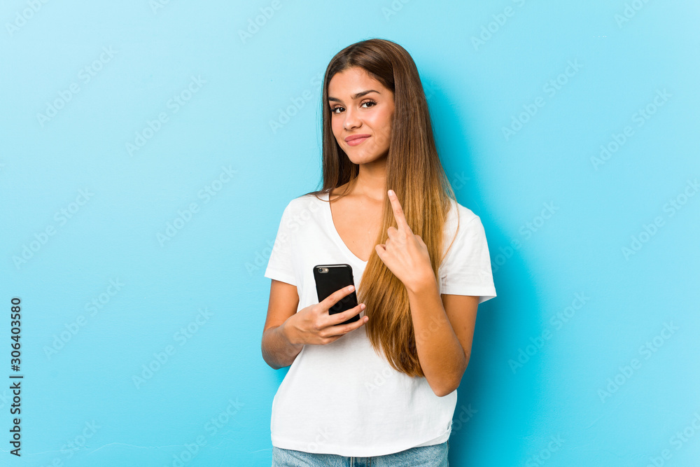 Young caucasian woman holding a phone pointing with finger at you as if inviting come closer. Young caucasian woman holding a phone pointing with finger at you as if inviting come closer.