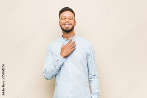 Young south-asian business man laughs out loudly keeping hand on chest.