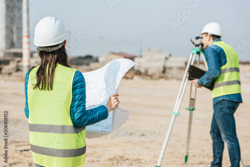 Selective focus of surveyor with blueprint and colleague using digital level on construction site
