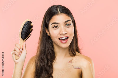 Young indian woman holding a hairbrush surprised pointing at himself, smiling broadly.