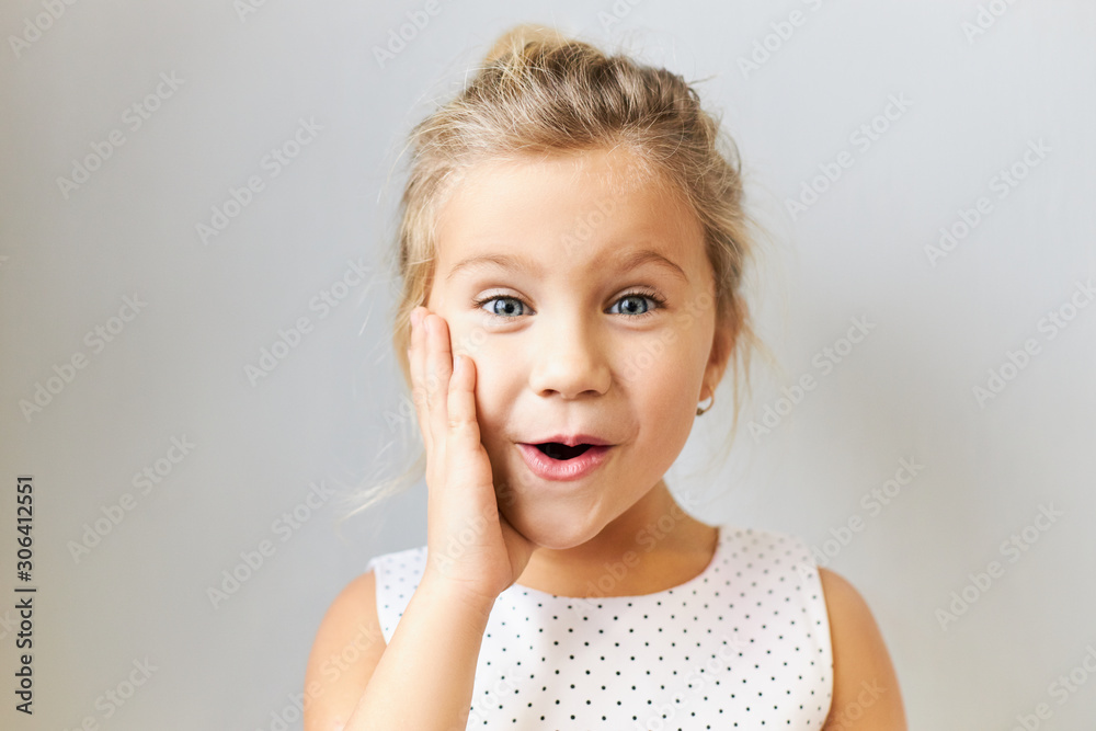 Adorable European girl of preschool age posing isolated with hand on her cheek, saying Wow with mouth opened, being amazed with exciting news, expressing true reaction, wearing dotted dress Adorable European girl of preschool age posing isolated with hand on her cheek, saying Wow with mouth opened, being amazed with exciting news, expressing true reaction, wearing dotted dress
