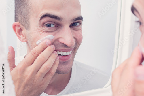 Skin care concept. Young man applying cream at his face and looking at himself with smile while standing in front of the mirror, close up, toned