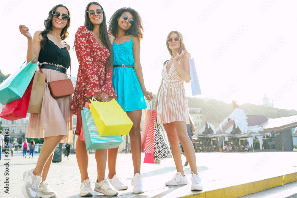 Summer sun. Four marvelous women in summer outfits are posing on the street in the city center, smiling with joy and showing their purchases to the camera. Summer sun. Four marvelous women in summer outfits are posing on the street in the city center, smiling with joy and showing their purchases to the camera.