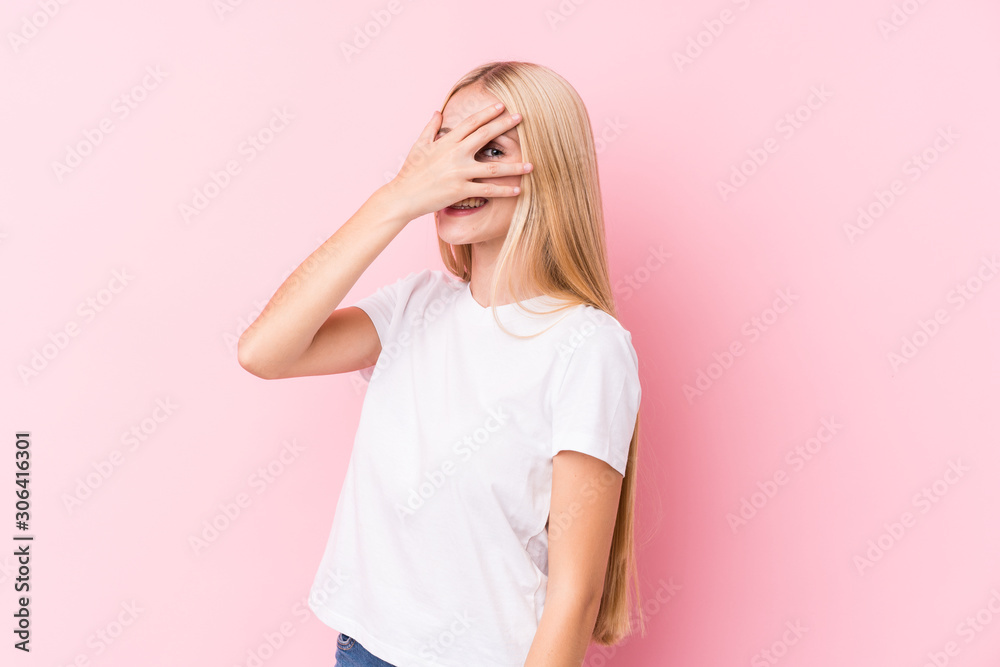 Young blonde woman on pink background blink at the camera through fingers, embarrassed covering face. Young blonde woman on pink background blink at the camera through fingers, embarrassed covering face.