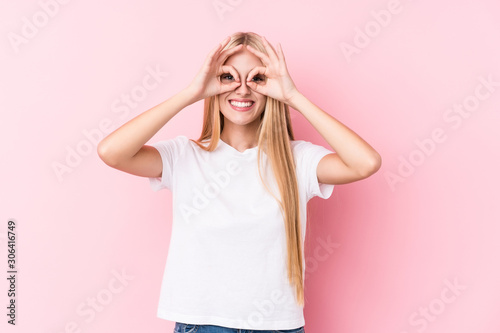 Young blonde woman on pink background showing okay sign over eyes