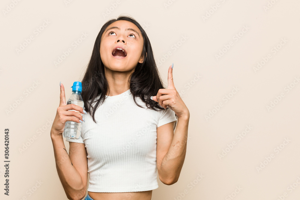 Young asian woman holding a water bottle pointing upside with opened mouth. Young asian woman holding a water bottle pointing upside with opened mouth.