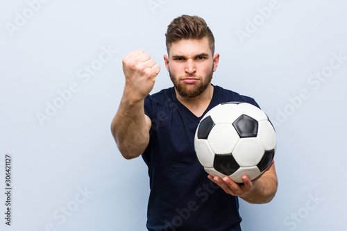 Young caucasian man holding a soccer ball showing fist to camera, aggressive facial expression.