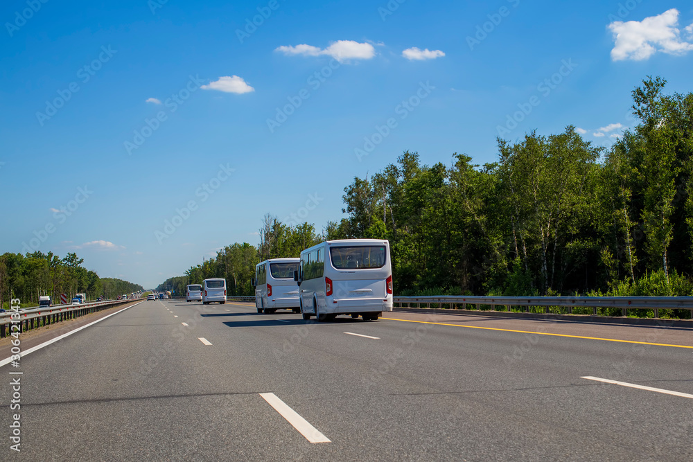 Four new white minibuses ride in a convoy along the highway on a sunny day in summer. back view