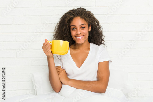 Young african american woman sitting on the bed holding a coffee mug smiling confident with crossed arms.