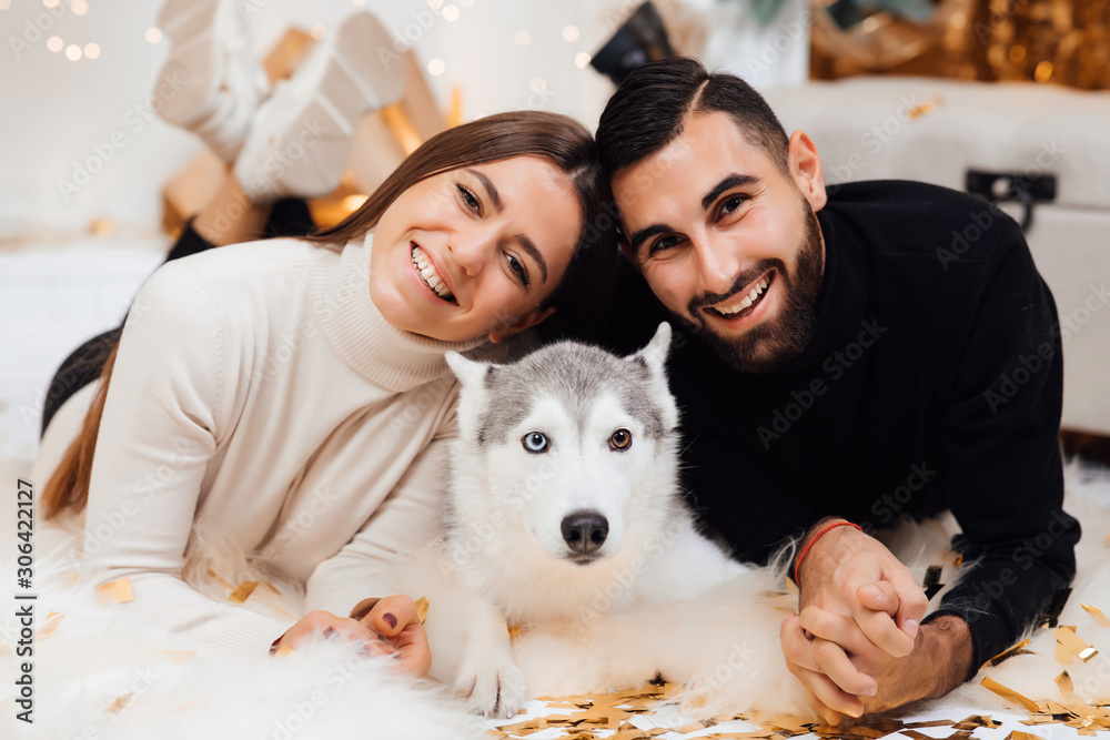 Beautiful young couple in the New Year atmosphere with dog of the Husky. Smiling couple enjoying in the holidays and playing with dog at home. Happy couple celebrating Christmas with their funny dog