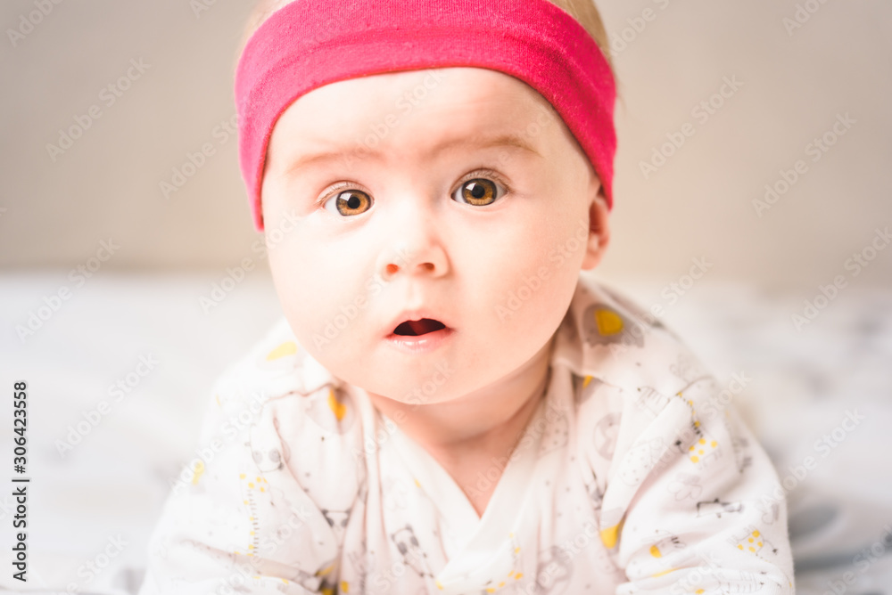 Adorable 6 months old Baby girl infant on a bed on her belly with head up looking into camera with her big eyes. Natural light. Adorable 6 months old Baby girl infant on a bed on her belly with head up looking into camera with her big eyes. Natural light.