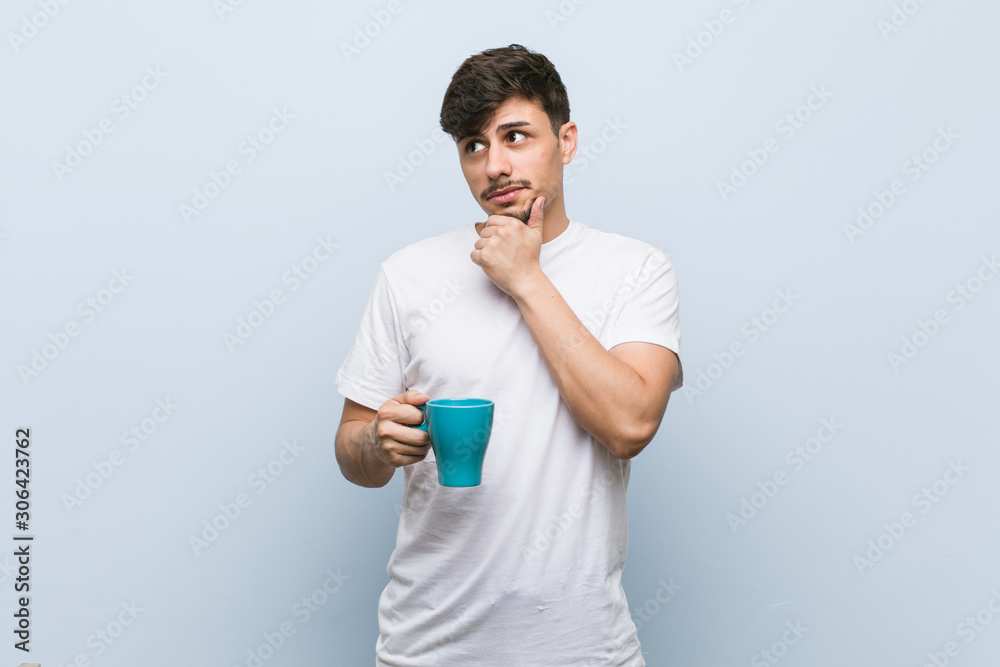 Young hispanic man holding a cup looking sideways with doubtful and skeptical expression. Young hispanic man holding a cup looking sideways with doubtful and skeptical expression.