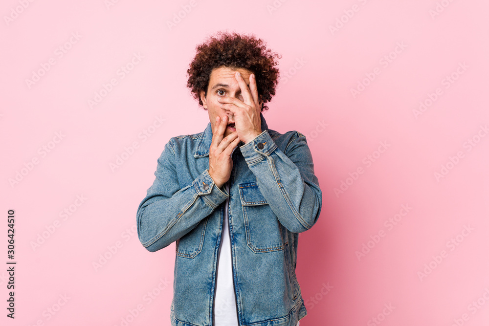 Curly mature man wearing a denim jacket against pink background blink through fingers frightened and nervous. Curly mature man wearing a denim jacket against pink background blink through fingers frightened and nervous.