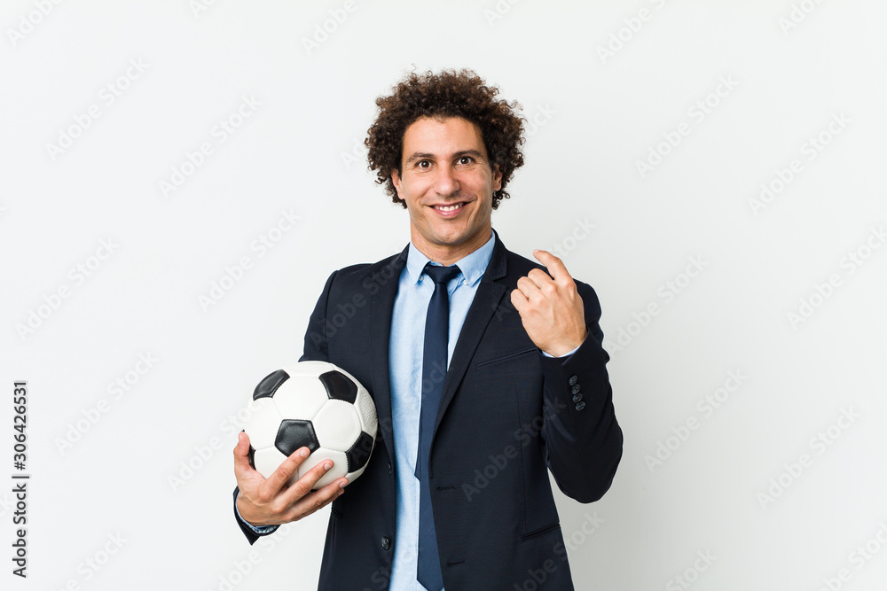 Soccer trainer holding a ball pointing with finger at you as if inviting come closer.