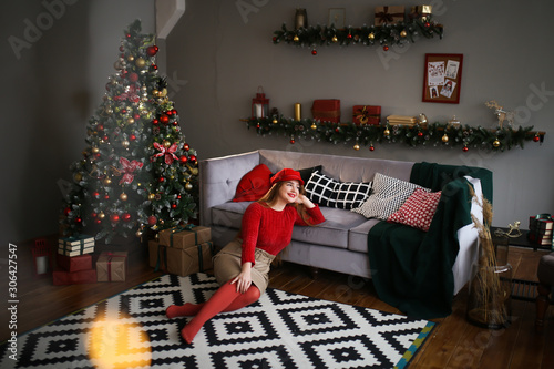 Beautiful bright attractive redhead girl plus size in a red sweater, beige leather skirt and red tights and a red cap rejoices and smiles on the background of the Christmas tree, gifts and bokeh.