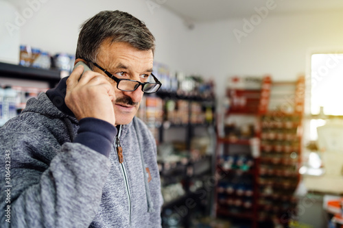 Senior man entrepreneur at his store talking to the phone making a call at work wearing eyeglasses