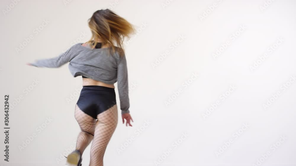 Studio shot of young energetic woman in fishnet tights and shorts performing dancehall and taking off cropped sweatshirt while getting undressed during dance against white background