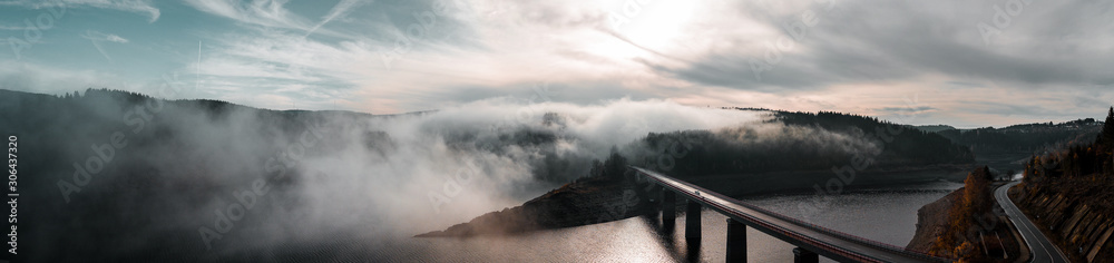 Fototapeta premium Aerial drone view from above of a morning mountain landscape with a lake dam and heavy low fog clouds hanging over the water and hills and winter mood.