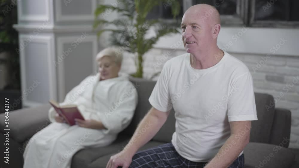 Concentrated senior bald man sitting on couch and watching TV when suddenly having heartache. His blond Caucasian wife putting aside book and coming to husband from background. Medicine, health, care. Concentrated senior bald man sitting on couch and watching TV when suddenly having heartache. His blond Caucasian wife putting aside book and coming to husband from background. Medicine, health, care.