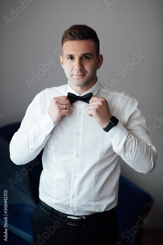 Grooms morning preparation, handsome groom straightens the bow tie and preparing for the wedding. Preparation of groom before wedding at home. Wedding morning groom.