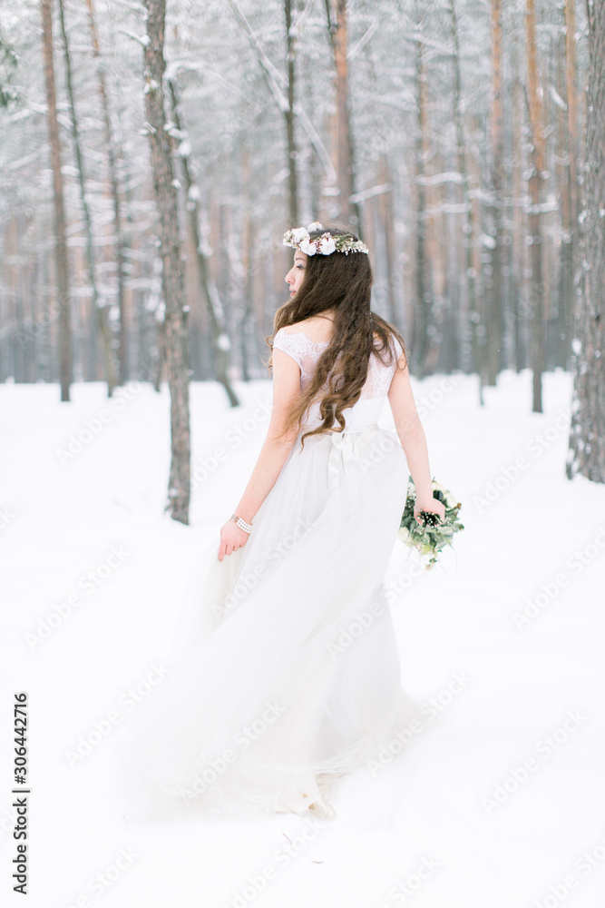 Back view of a beautiful young woman, bride, walking in a winter forest. Bride with wreath on the head, with long curly hair, holding winter bouquet