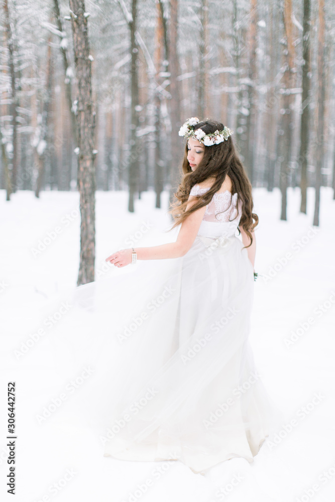 Back view of a beautiful young woman, bride, walking in a winter forest. Bride with wreath on the head, with long curly hair, holding winter bouquet Back view of a beautiful young woman, bride, walking in a winter forest. Bride with wreath on the head, with long curly hair, holding winter bouquet