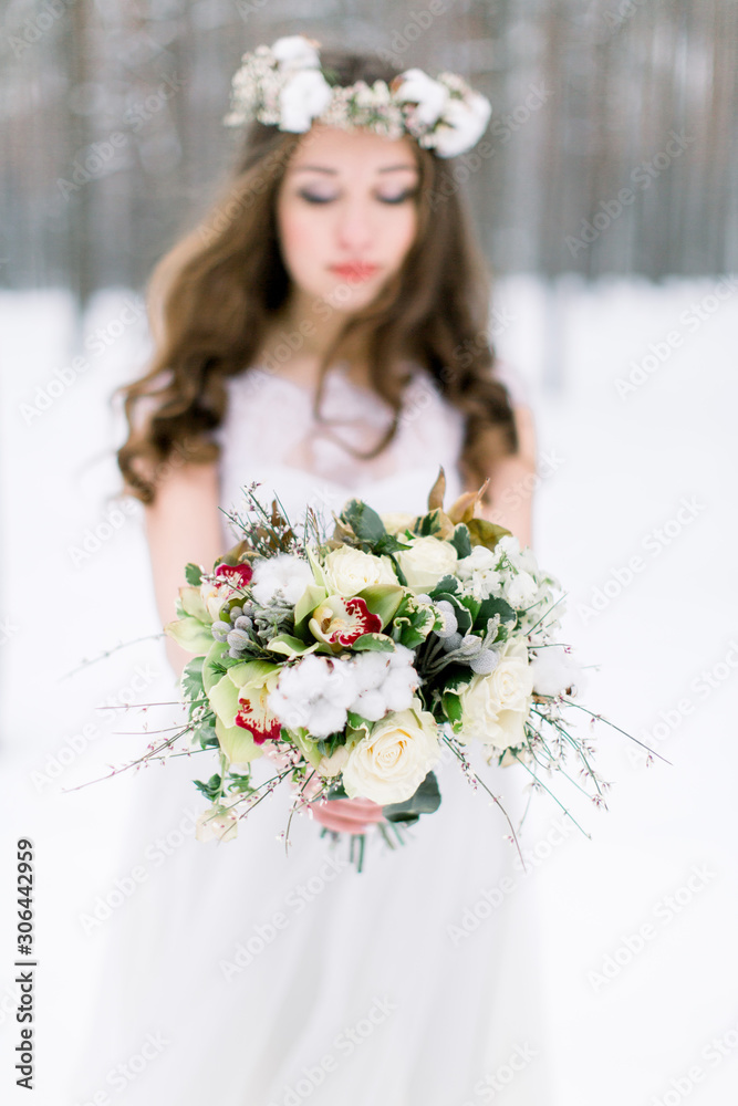 Beautiful bride. Winter wedding. Young woman in wedding dress with a floral cotton head wreath and bouquet standing in the winter forest and looking at the camera Beautiful bride. Winter wedding. Young woman in wedding dress with a floral cotton head wreath and bouquet standing in the winter forest and looking at the camera