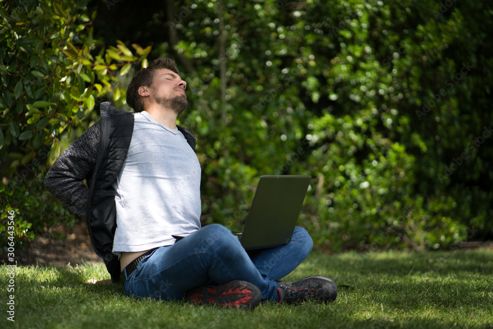 Young caucasian man sitting on the grass of a park with his computer. He is complaining about his back pain. Natural Environment Young caucasian man sitting on the grass of a park with his computer. He is complaining about his back pain. Natural Environment