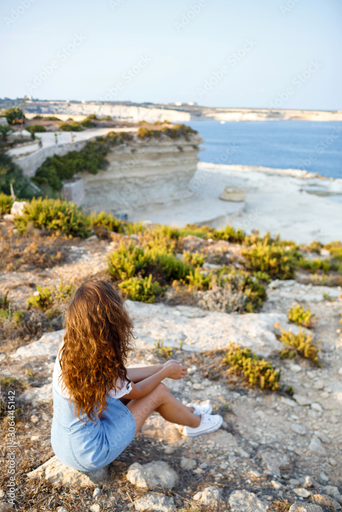 A slender female tourist sits on a cliff at a height and considers the beauty of nature, the sea and rocks. Back view.