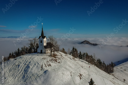 Church of Sveti Jakob standing on the top of the hill surrounded by clouds in the valley on a sunny winter day
